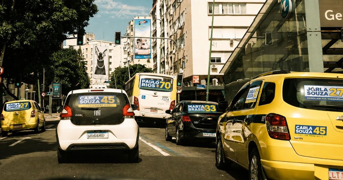 Carros e ônibus no trânsito de uma avenida urbana com adesivos de campanha eleitoral colados nos vidros e para-choques